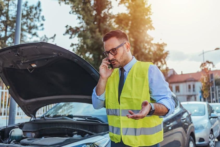 Egy láthatósági mellényt viselő férfi egy nyitott autó motorháztetője mellett áll.