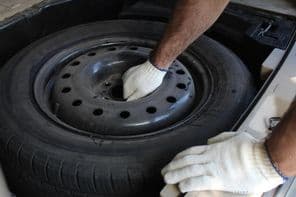 Egy középkorú férfi fehér munkakesztyűt visel, és kiszed egy pótkereket a csomagtartóból.