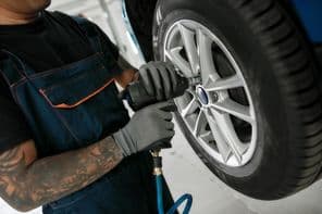 Egy férfi szerelő, aki pneumatikus kulccsal csavarozza le egy felemelt autó kerekét.