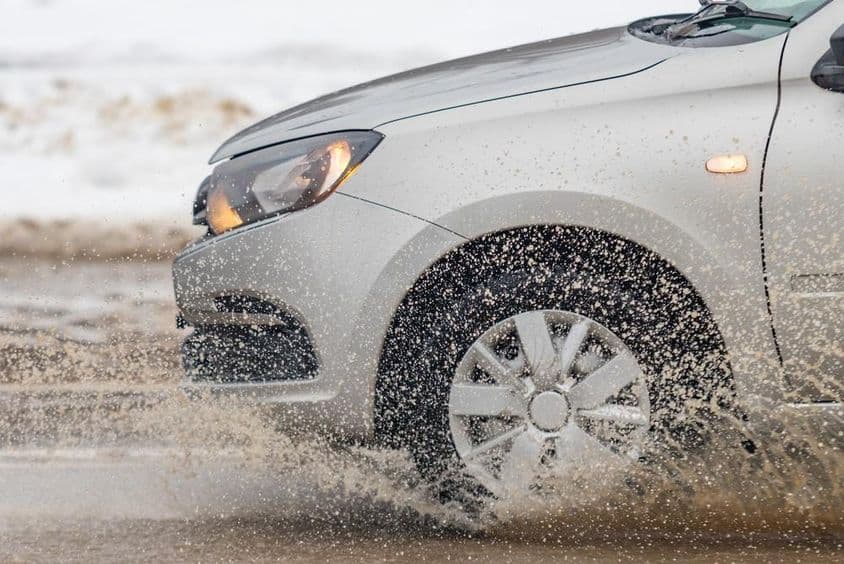 Egy autó sáros úton halad, bekapcsolt fényszórókkal.